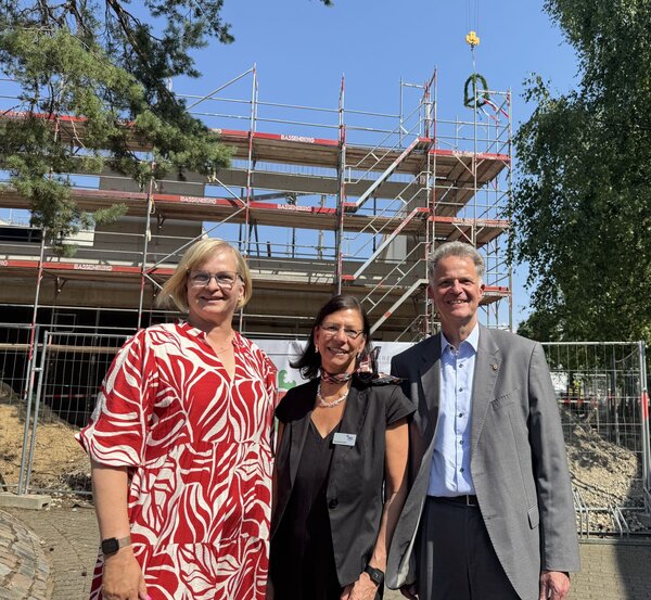 Zwei Frauen und ein Mann stehen vor einer Baustelle mit Gerüst. Im Hintergrund hängt eine grüne Richtfestkrone an einem Kranseil.