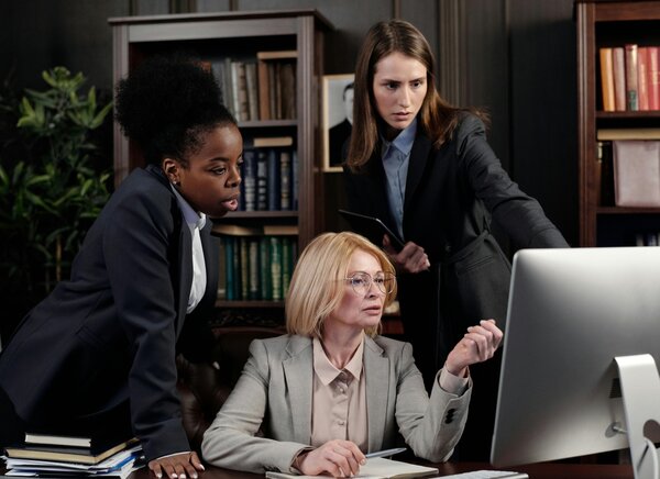 Drei Frauen stehen und sitzen vor einem Schreibtisch mit einem Computer.