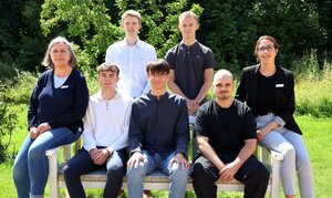 Zwei Frauen und fünf junge Männer posieren auf und hinter einer Holzbank im Freien.