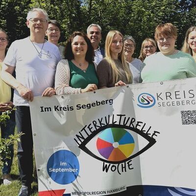 Frauen und Männer stehen in einem grünen Garten und halten ein großes Plakat der Interkulturellen Woche hoch.