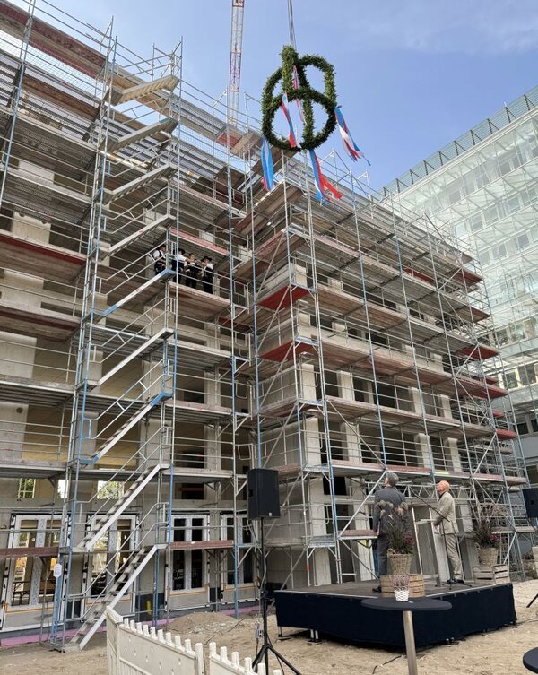 Der Landrat und ein Architekt stehen auf der Bühne vor einem Rohbau und sehen nach oben. Dort ist die Richtkrone zu sehen. Drei Zimmerer stehen in der vierten Etage und sprechen den Richtspruch.