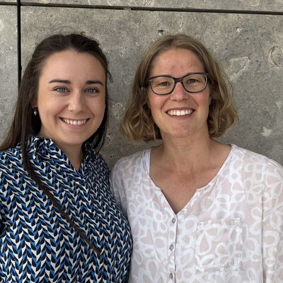 Zwei Frauen stehen vor einer Steinwand und lachen in die Kamera.