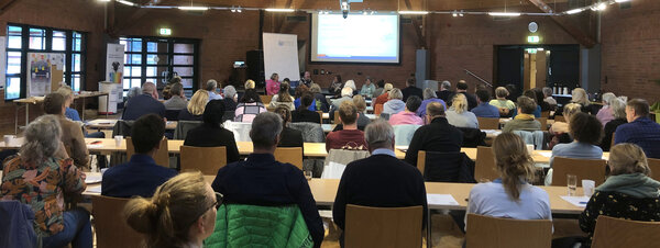 In einem großen Raum sind Menschen von hinten zu sehen, die auf Stühlen an langen Tischen sitzen und nach vorne schauen.