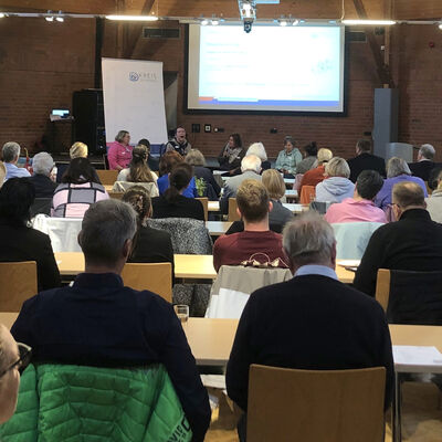 In einem großen Raum sind Menschen von hinten zu sehen, die auf Stühlen an langen Tischen sitzen und nach vorne schauen.