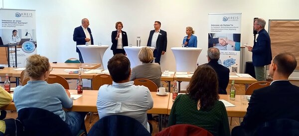 Vier Personen stehen auf einem Podium und sprechen miteinander. Davor sitzt das Publikum.