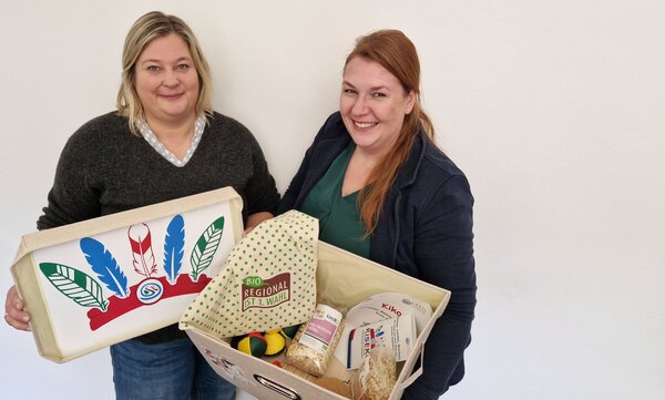 Zwei Frauen lachen in die Kamera. Eine hält eine Box in der Hand, die andere den Deckel. In der Box befinden sich unter anderem Müsli, Stoffbälle und Informationsbroschüren.