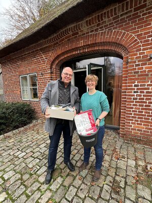 Ein Mann und eine Frau stehen vor einem Reetdachhaus und halten eine Pappbox beziehungsweise einen Eimer mit Arbeitsmaterialien in den Händen.
