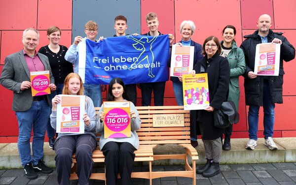 Schüler*innen und Erwachsene halten Plakate und Flaggen in die Kamera. Es geht um Gewalt gegen Frauen. Auf einer Holzbank steht "Kein Platz für Gewalt".