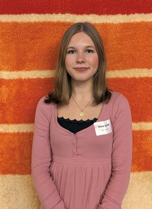 Eine jugendliche Schülerin steht vor einem bunten Wandteppich und lächelt in die Kamera.