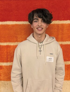 Ein dunkelhaariger jugendlicher Schüler steht vor einem Wandteppich und lächelt in die Kamera.