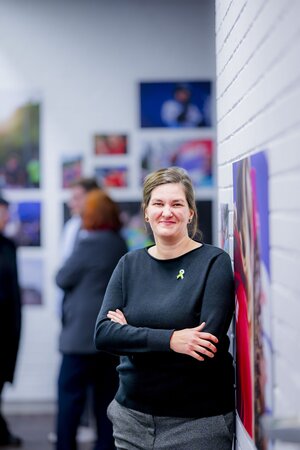 Eine Frau lehnt an einer Wand und lächelt. An der Seite und im Hintergrund sind verschwommene, großformatige Fotografien zu erkennen.