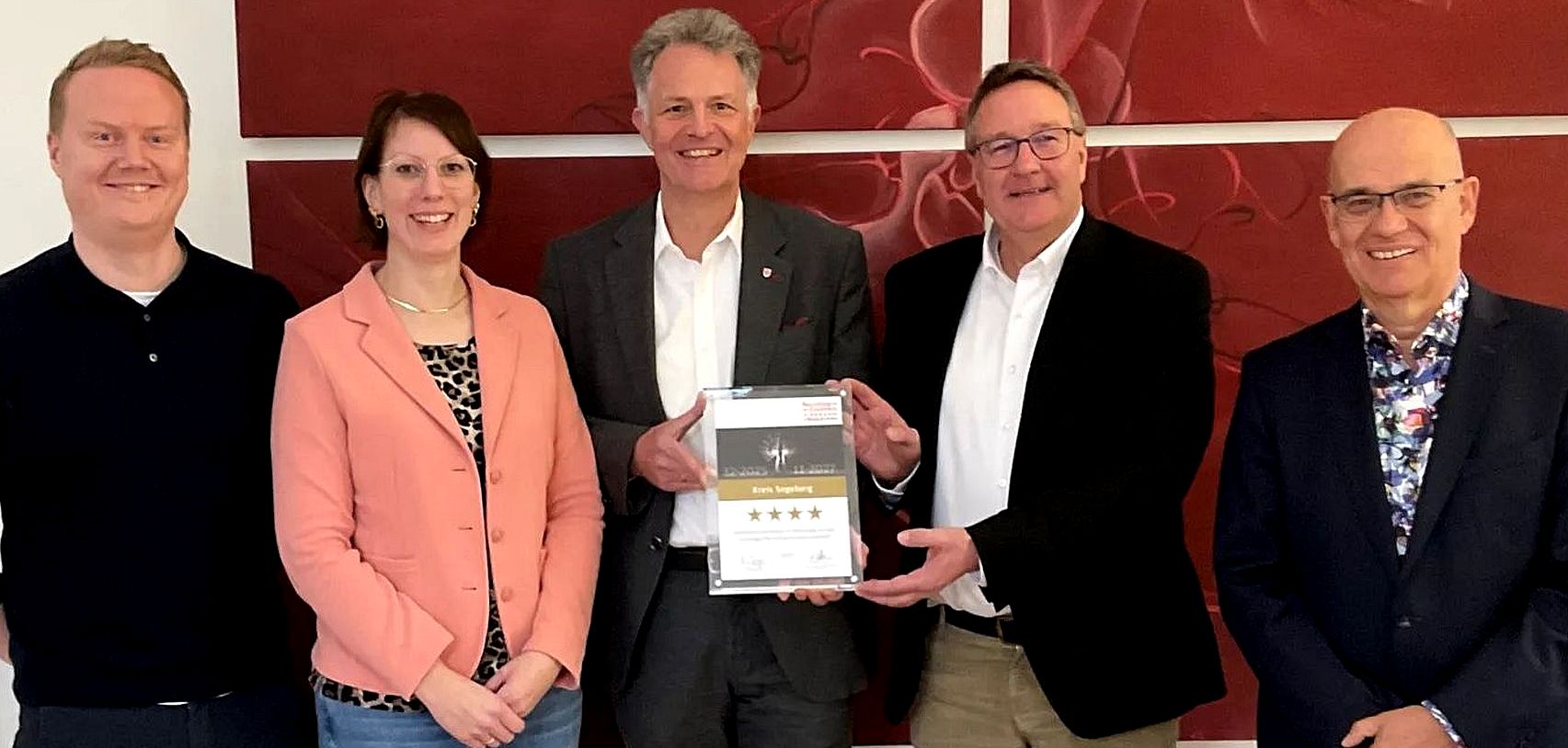 Drei M&auml;nner und eine Frau stehen vor einer Wand und l&auml;cheln in die Kamera. zwei der M&auml;nner halten eine Auszeichnung in der Hand.