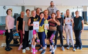 Mehrere Frauen und ein Mann stehen in Sportkleidung in einem Fitnessraum und l&auml;cheln.