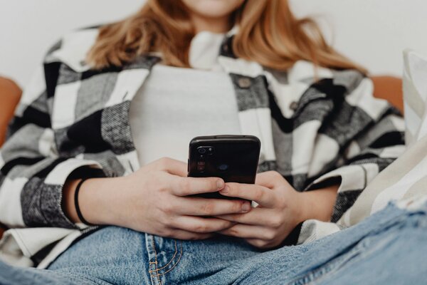Eine Person mit langen Haaren sitzt auf einem Sofa und h&auml;lt ein Handy in der Hand. Das Gesicht ist nicht zu erkennen/abgeschnitten.