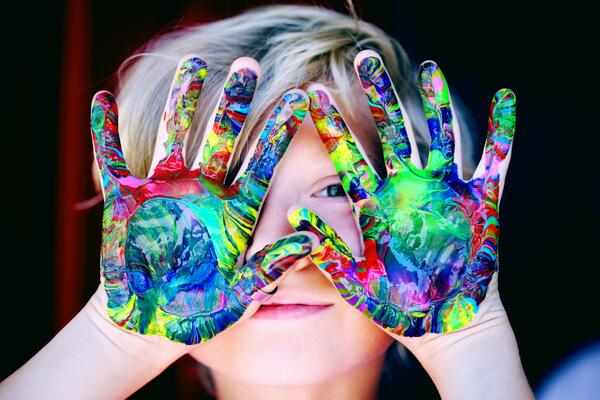Ein Kind hält bunt angemalte Hände vor das Gesicht und schaut zwischen den Fingern hindurch.