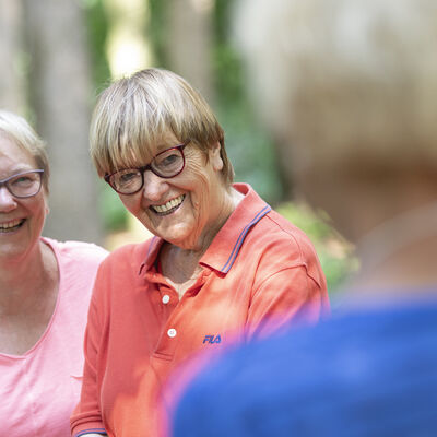 Drei ältere Damen stehen im Wald und lächeln einander an.