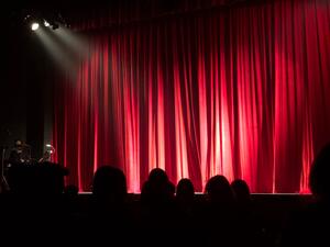 Ein roter Vorhang auf einer Theaterbühne. Im Vordergrund sieht man die Silhouetten der Zuschauer*innen.