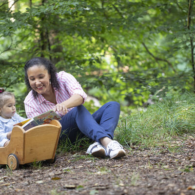 Mutter liest einem kleinen Kind im Wald vor. Das Kind sitzt in einem Holzpuppenwagen.