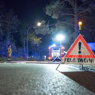 Ein Feuerwehrauto steht mit ausgerollten Schläuchen bei Nacht auf einer Straße. Darum stehen viele Bäume. In einiger Entfernung vor dem Feuerwehrauto steht ein Wahndreieck mit der Aufschrift "Feuerwehr".