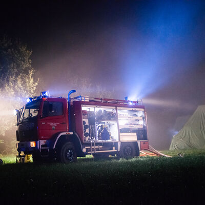 Ein Feuerwehrauto steht im Dunkeln auf einem Feld. Daneben sind Bäume und Zelte zu sehen.