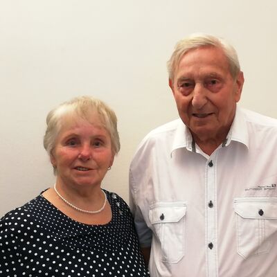 Ein Foto von einem Mann und einer Frau vor einer weißen Wand. Der Mann trägt ein weißes Hemd. Die Frau trägt ein schwarzes Oberteil mit weißen Punkten.