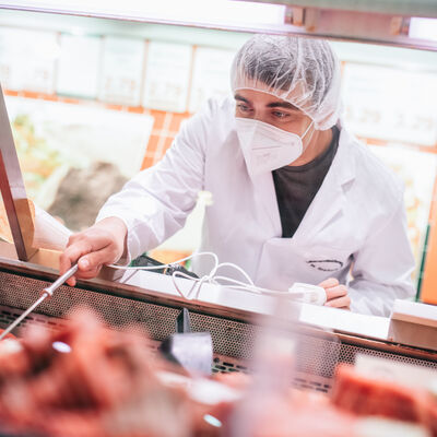 Ein Mitarbeiter steht im Kittel mit Mundschutz und Haarnetz an einer Fleischtheke. Er überprüft die Temperatur des Fleisches.