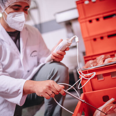 In vielen roten Kisten ist Fleisch gelagert. Ein Mitarbeiter im Kittel mit Mundschutz und Haarnetz überprüft die Qualität des Fleisches.