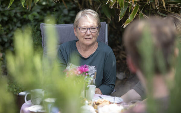 Eine Frau sitzt mit mehreren Personen am Kaffeetisch. Im Hintergrund sind grüne Büsche zu sehen.