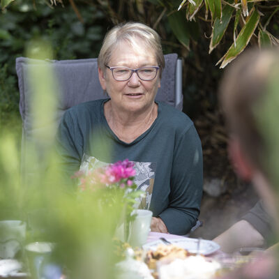 Eine Frau sitzt mit mehreren Personen am Kaffeetisch. Im Hintergrund sind grüne Büsche zu sehen.