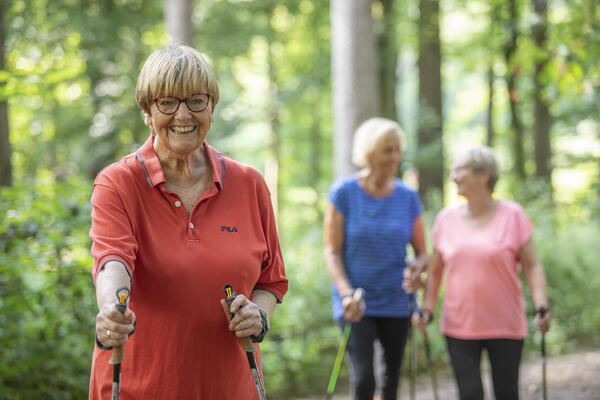 Drei ältere Damen laufen sportlich mit Nordic-Walking-Stöcken durch den Wald.
