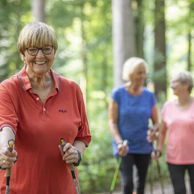 Drei ältere Damen laufen sportlich mit Nordic-Walking-Stöcken durch den Wald.