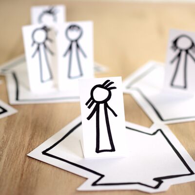 Gemalte Strichmnnchen auf Papier stehen ausgeschnitten und aufrecht auf gemalten Husern, die flach auf einem Holztisch liegen.